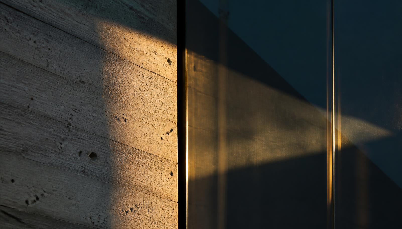 Detalhe de parede de concreto aparente encontrando um painel de vidro escuro sob luz rasante dourada
