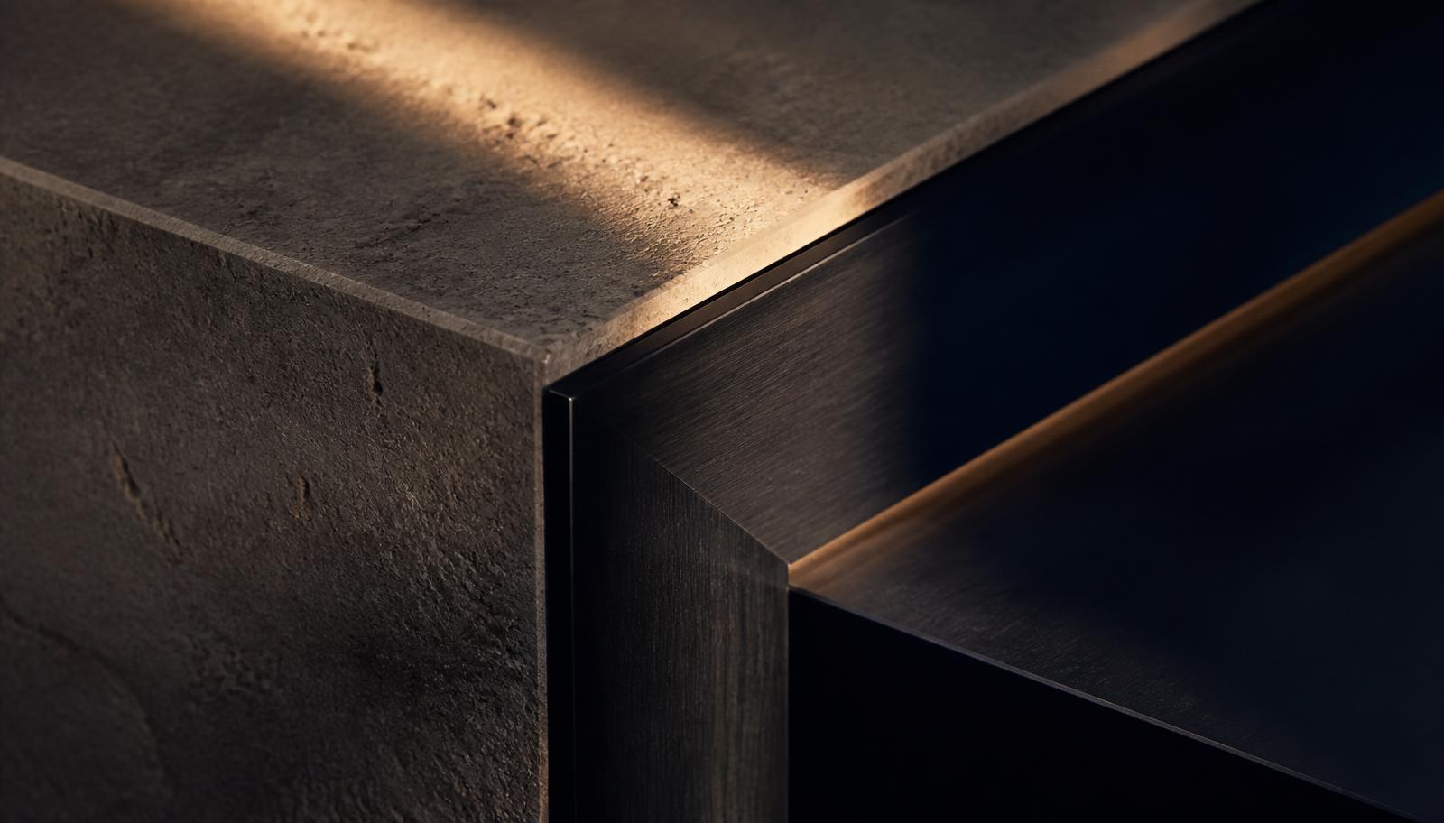 Detalhe de encontro entre pedra natural e metal escovado escuro sob luz dourada rasante